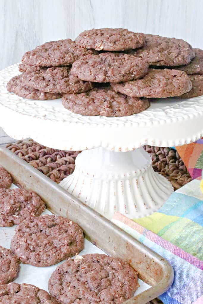 A vertical image of a cake plate filled with Chocolate Oatmeal Cookies along with a baking sheet filled with cookies and a colorful napkin.
