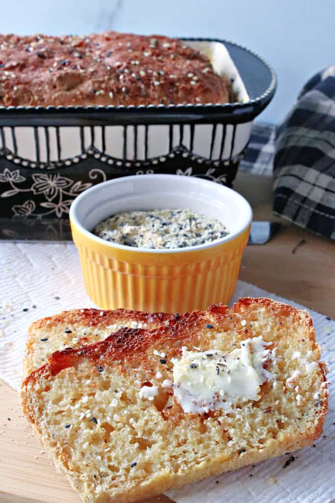 A vertical photo of a few slices of Everything English Muffin Bread in the forefront along with an entire loaf in the background.