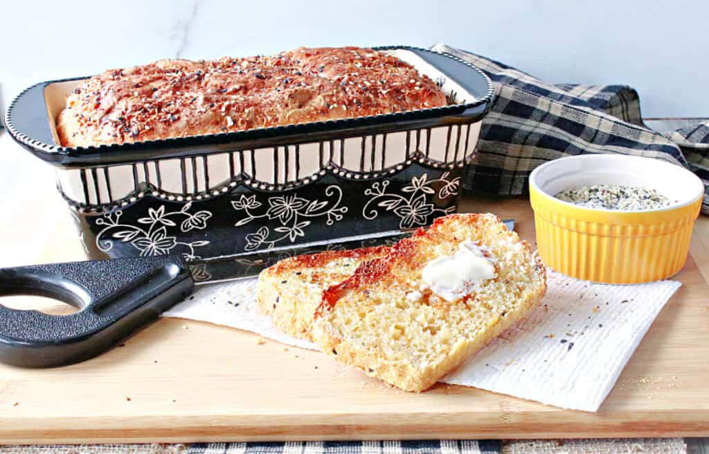 A horizontal photo of a loaf of Everything English Muffin Bread in a black and white loaf pan along with some black and tan checkered napkins and a knife. 