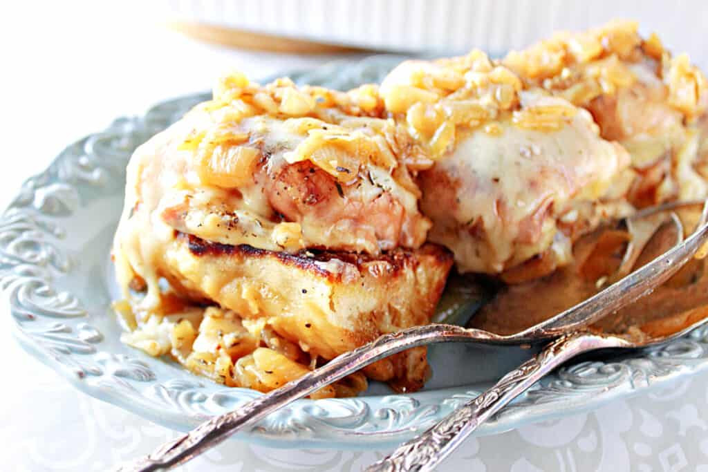 A few French Onion Chicken Thighs on a pretty blue plate with silver serving utensils, melted cheese, and caramelized onions.