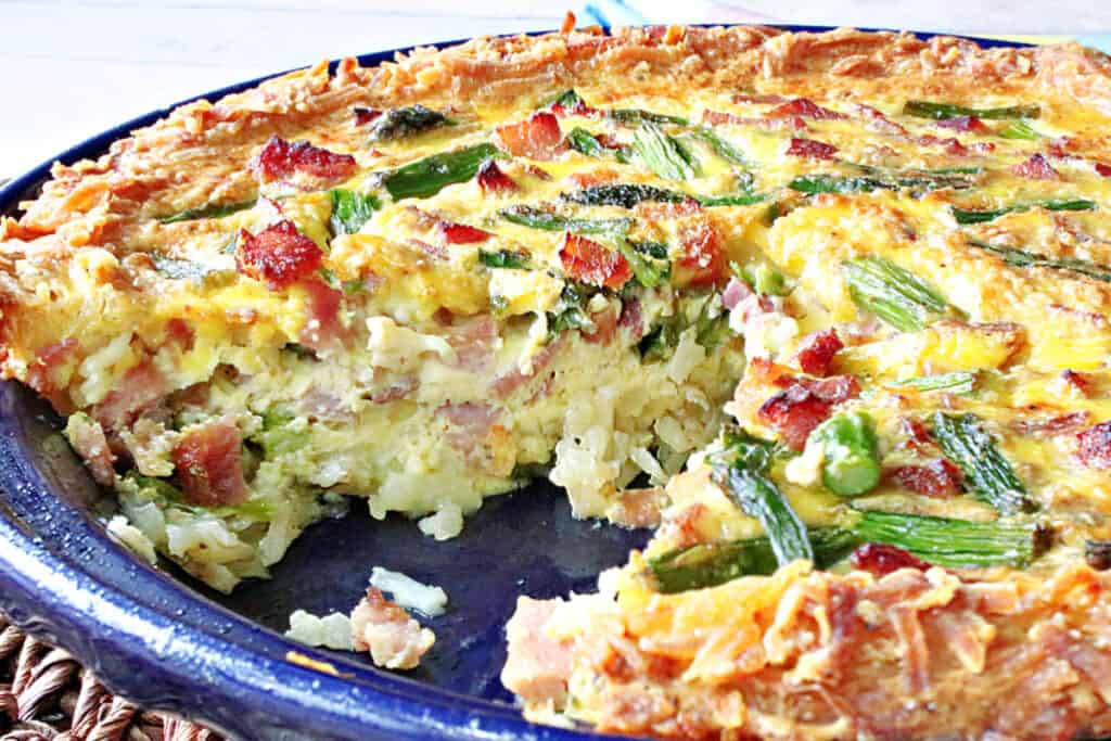 A horizontal closeup photo of a Ham and Asparagus Quiche with a slice taken out.