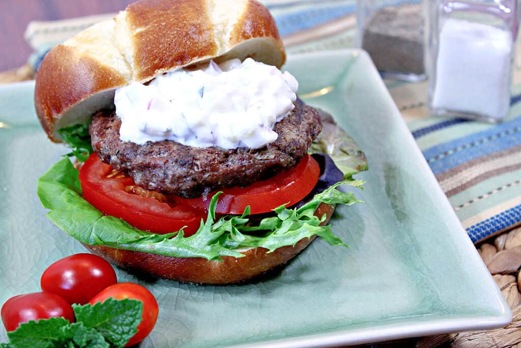 An offset horizontal photo of a Lamb Burgers with Feta and Tzatziki sauce on a green plate with tomatoes, lettuce, and a bun.