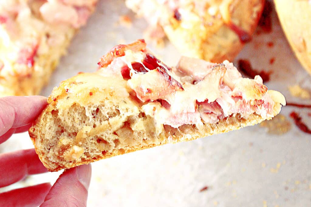 A hand holding a slice of Leek and Onion Focaccia with ham and melted cheese. 