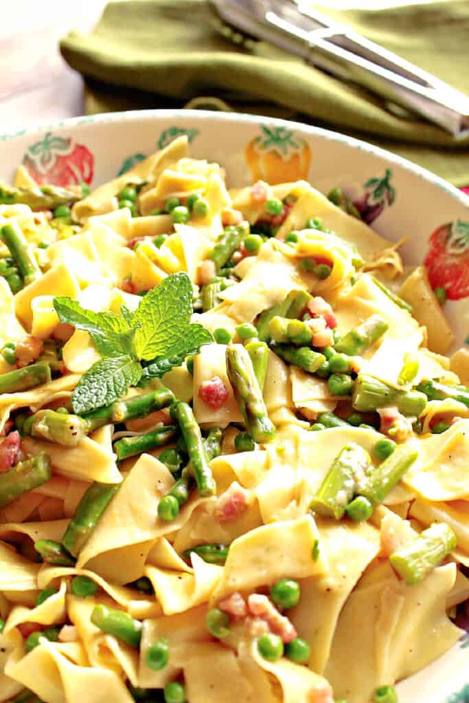 A closeup vertical image of Pappardelle with Spring Vegetables in a ceramic bowl with fresh mint, peas, asparagus, and pancetta.