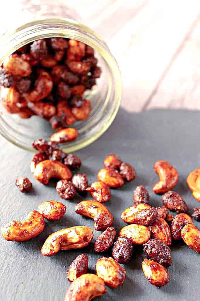 A vertical closeup image of Brown Butter Roasted Nuts spilled out onto a black slate tile with a tipped jar in the background.