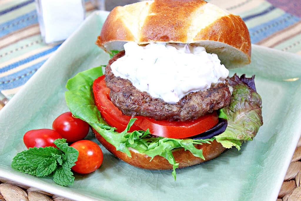 A Greek Lamb Burger with Feta and Tzatziki on a green square plate with tomato and lettuce.