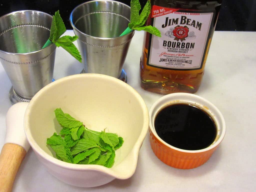 A horizontal set up of the making of Traditional Mint Juleps along with metal cups, a mortar and pestle, and a bottle of Kentucky bourbon.