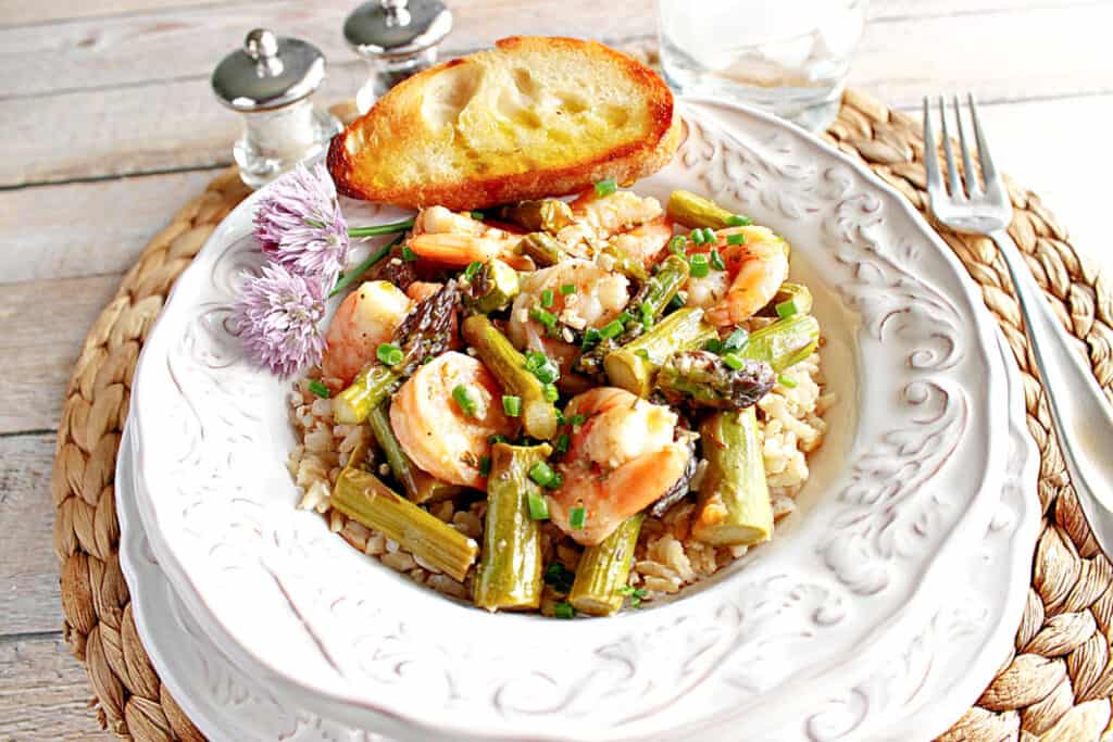 A pretty white bowl filled with Shrimp and Asparagus along with chives, garlic bread, and clover flowers.