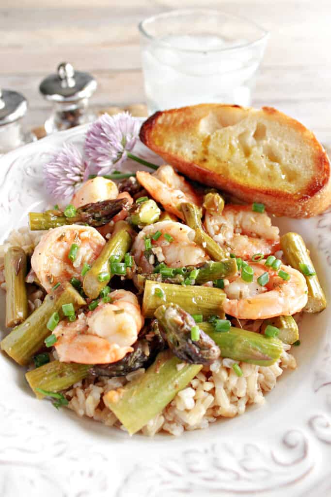 A vertical closeup image of Shrimp and Asparagus in a bowl with a piece of garlic bread and some clover flowers.