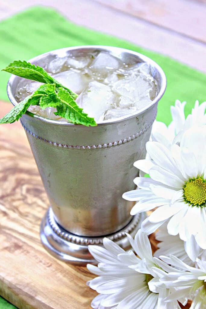 A vertical closeup photo of a Traditional Mint Julep in a sliver metal cup along with ice and fresh mint leaves.