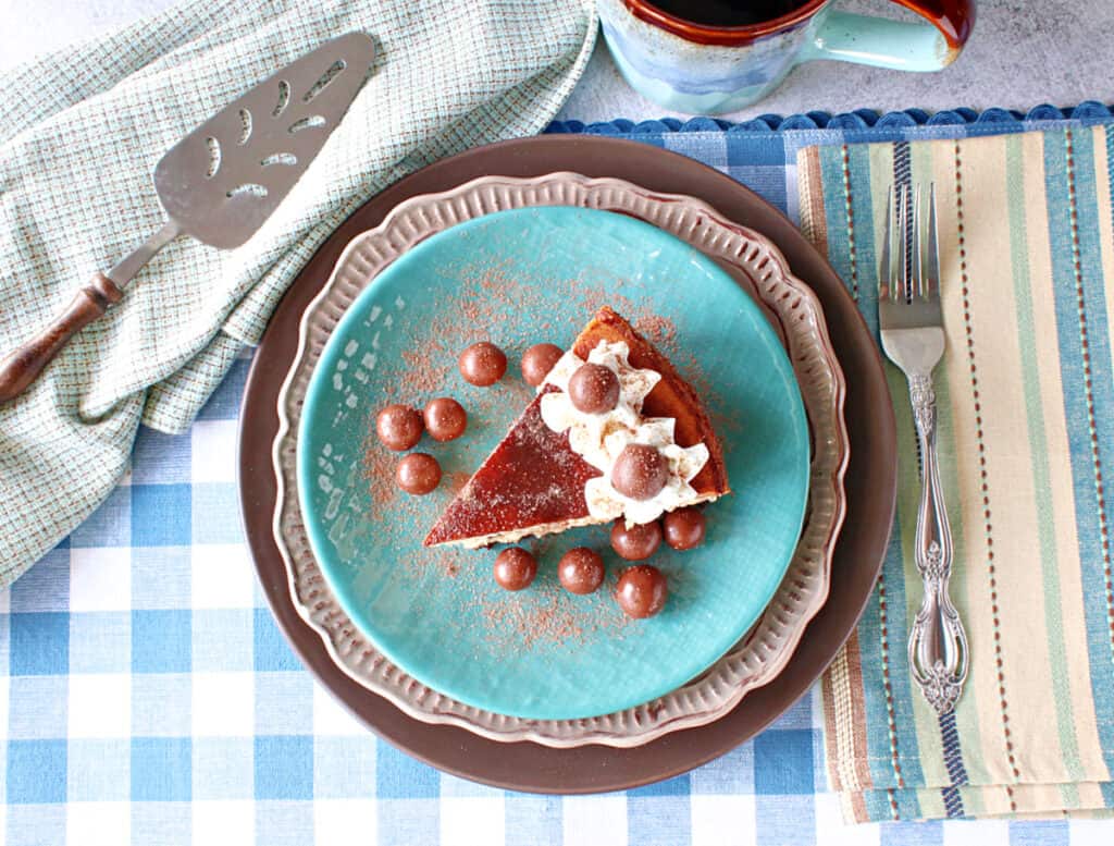 A direct overhead photo of a slice of Malted Milk Ball Cheesecake on a blue plate with a fork on the side along with a cake server.