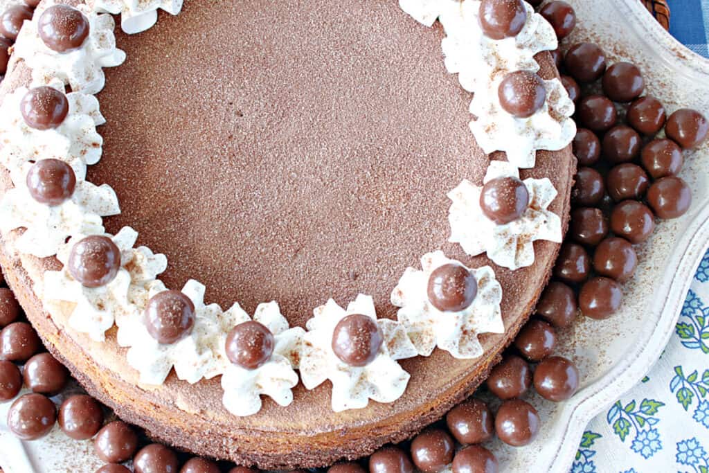 An overhead closeup offset photo of a Malted Milk Ball Cheesecake with whipped cream garnish and malted milk ball candy accents.