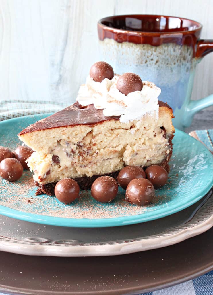 A slice of Malted Milk Ball Cheesecake on a blue plate surrounded with malted milk balls and topped with whipped cream.