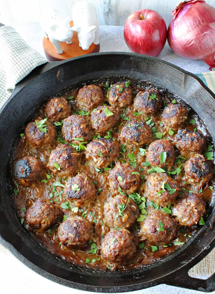 A vertical overhead closeup of Pork Meatballs with Apple and Onion in a cast iron skillet along with a fresh apple and red onion in the background.