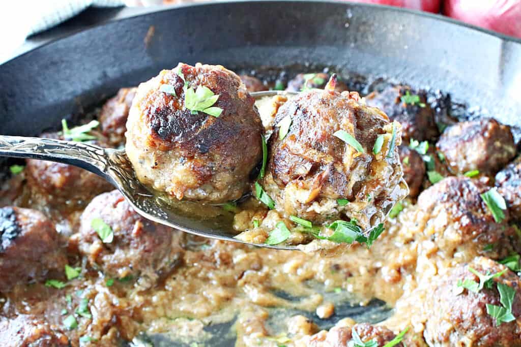 A serving spoon holding two Pork Meatballs with Apple and Onion over a cast iron skillet filled with meatballs. 