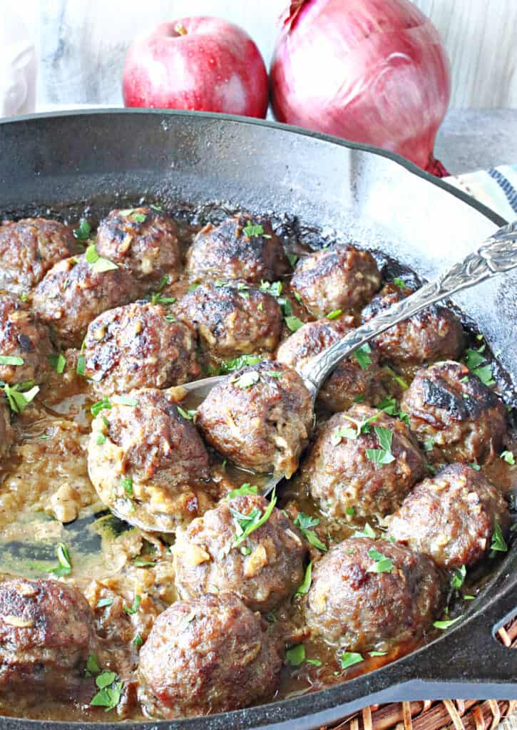 A vertical closeup of a serving spoon in a cast iron skillet holding two Pork Meatballs with Apple and Onion along with fresh parsley.