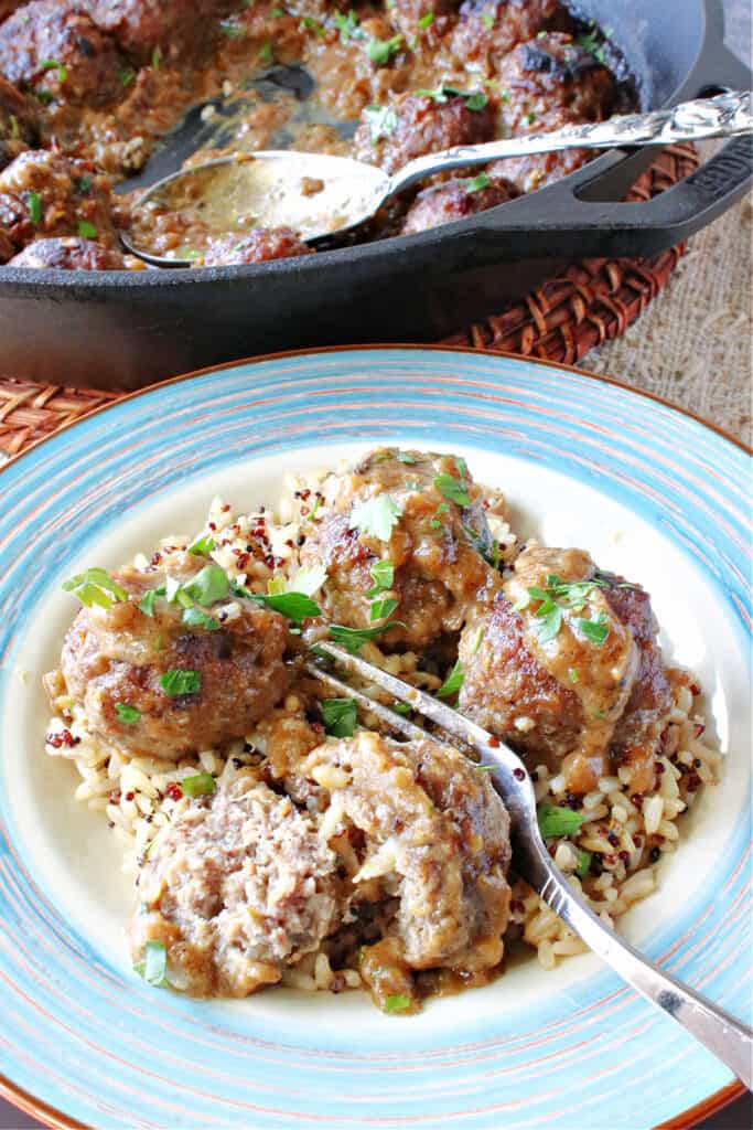 A vertical closeup image of a plate filled with Pork Meatballs with Apple and Onion in the foreground and a skillet with a serving spoon in the background.