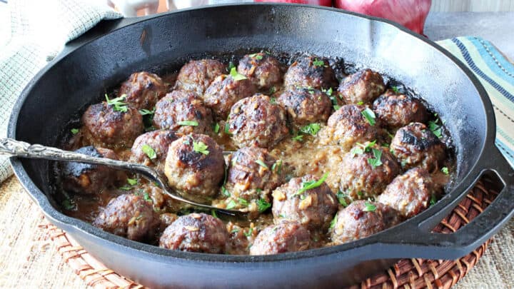 A cast iron skillet filled with Pork Meatballs with Apple and Onion and a serving spoon.