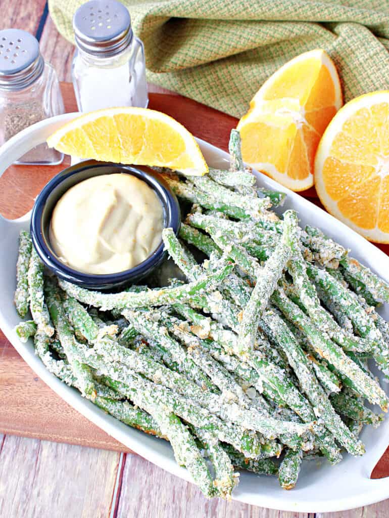 A vertical overhead closeup of a bowl of Everything Green Bean Fries along with a small round dish of Orange Honey Dipping Sauce and fresh oranges.