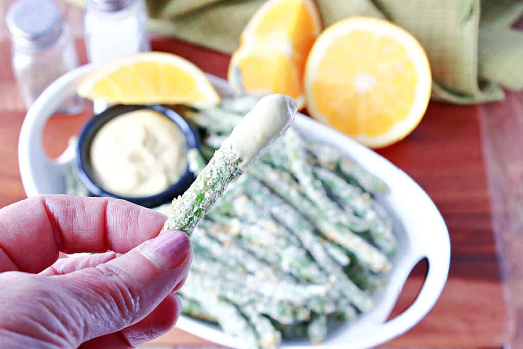 A closeup horizontal photo of a hand holding an Everything Green Bean Fry that's been dipped into Orange Honey Dipping Sauce.