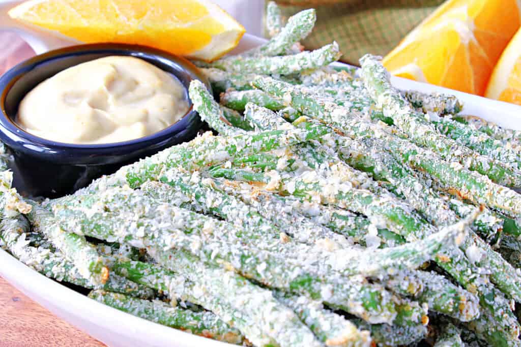 A horizontal closeup photo of a bowl filled with Everything Green Bean Fries covered in instant potato flakes along with an orange honey dipping sauce.