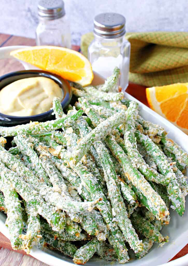 A vertical closeup of a dish of Everything Green Bean Fries along with s small dish of Orange Honey Dipping Sauce, orange wedges, and a salt and pepper shaker.