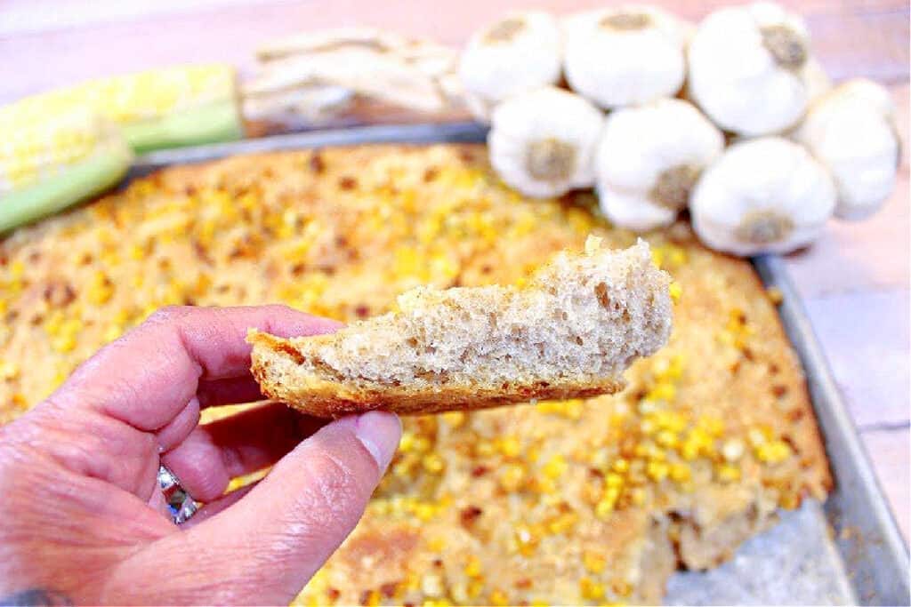 A horizontal photo of a closeup of a piece of Garlic Focaccia with Sweet Corn ripped from the full focaccia.