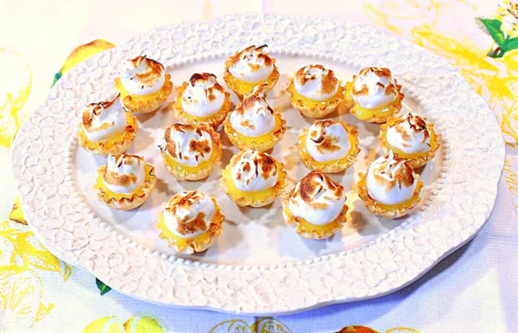 An overhead horizontal photo of a platter of Lemon Meringue Tartlets along with a lemon patterned napkin.