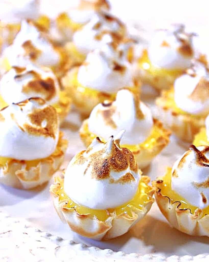 A closeup image of Lemon Meringue Tartlets on a white platter with toasted meringue topping.