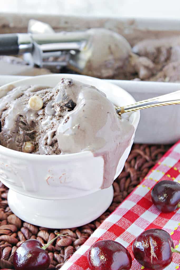 A vertical closeup image of a melting dish of Chocolate Cherry Kahlua Ice Cream with fresh cherries in the front and a red and white napkin.