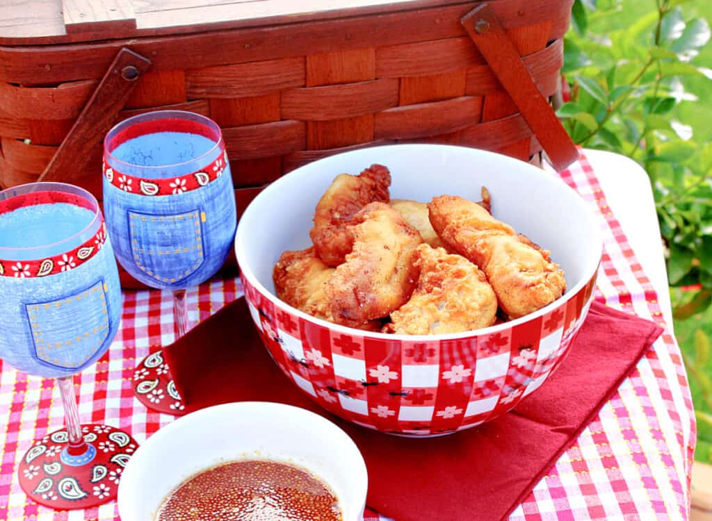 A picnic table set with a bowl of Waffle Batter Fried Chicken Tenders along with two glasses of wine and a picnic basket.