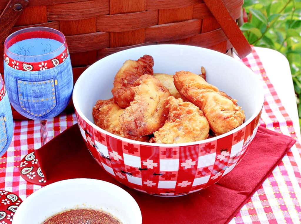 A closeup horizontal photo of a red and white bowl filled with Waffle Batter Fried Chicken Tenders outside on a picnic table.
