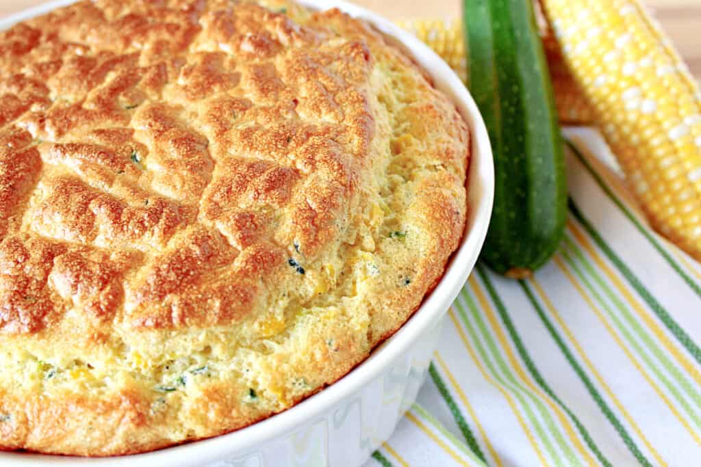 An offset horizontal photo of a Zucchini and Sweet Corn Soufflé in a white casserole dish with a zucchini and sweet corn in the background.