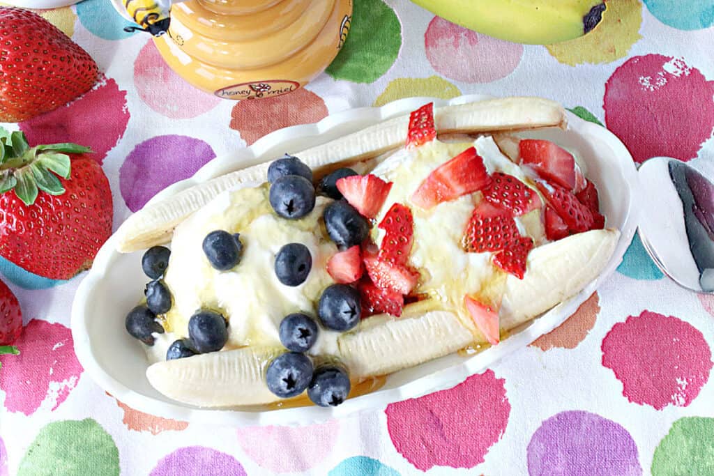 A direct overhead photo of a Frozen Greek Yogurt Banana Split in a dish with a fresh banana, honey, strawberries, and blueberries.