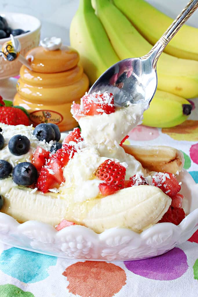 A vertical closeup of a spoon taking a scoop out of a Frozen Greek Yogurt Banana Split with fresh strawberries and blueberries on top.