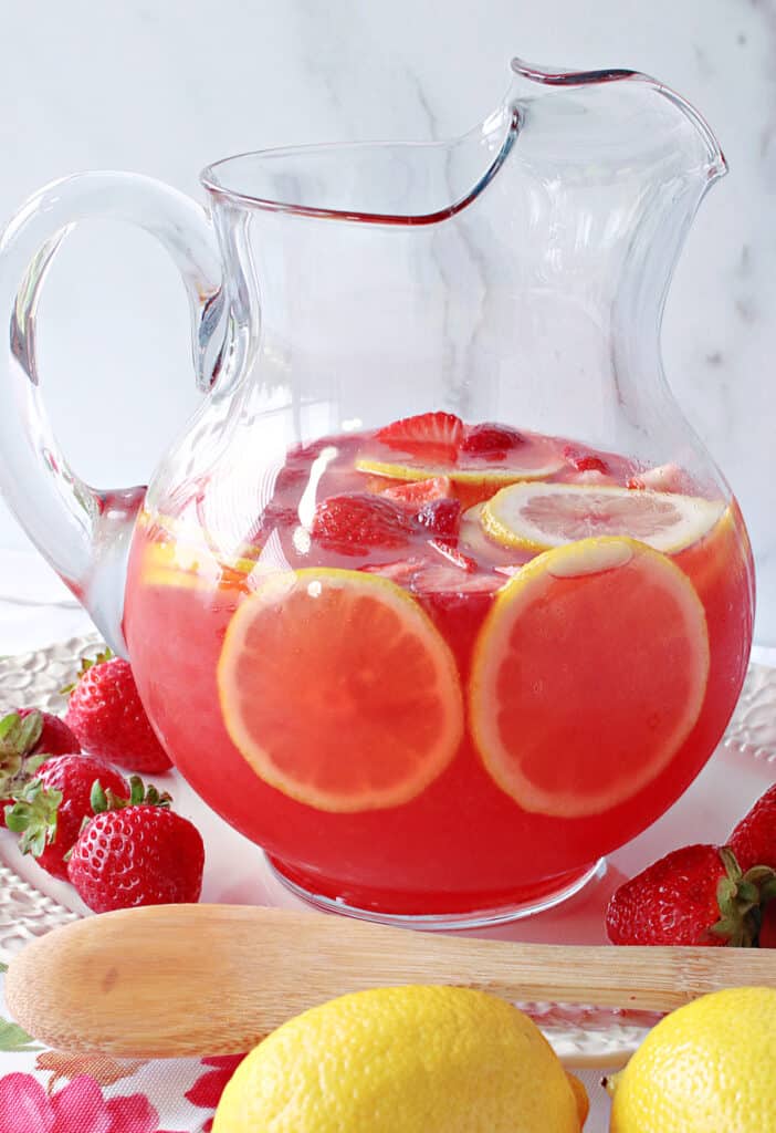 A vertical closeup of a pitcher filled with Lite Pink Lemonade with Strawberry Simple Syrup along with some fresh lemons, strawberries and a wooden spoon.