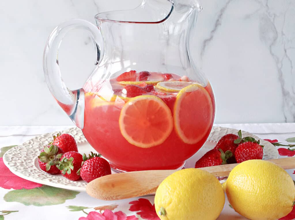 A horizontal photo of a glass pitcher filled with Lite Pink Lemonade with Strawberry Simple Syrup on a pretty platter with fresh lemons and strawberries on the platter.