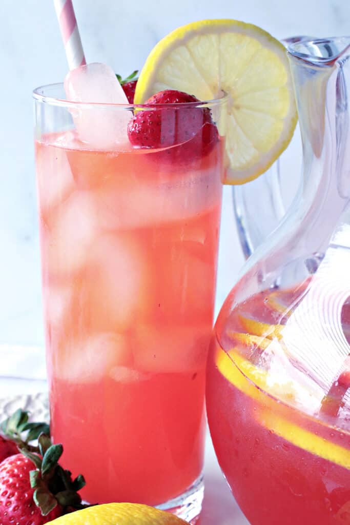A closeup vertical photo of a tall glass of Lite Pink Lemonade with Strawberry Simple Syrup along with a lemon slice and a strawberry in the glass.