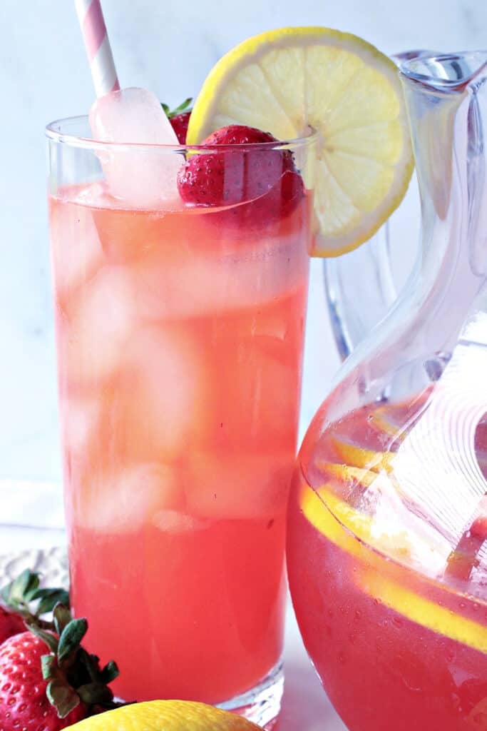 A tall glass of Lite Pink Lemonade with Strawberry Simple Syrup along with a lemon slice and a strawberry in the glass.