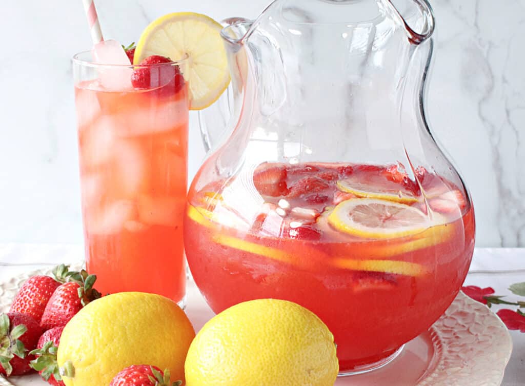 A horizontal photo of a glass and a glass pitcher filled with Lite Pink Lemonade with Strawberry Simple Syrup along with fresh lemons and strawberries in the photo as garnish.