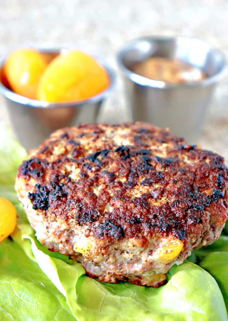 A closeup vertical image of a Pork Burger with Dried Apricots on a lettuce leaf along with two containers in the background with apricots and mustard.