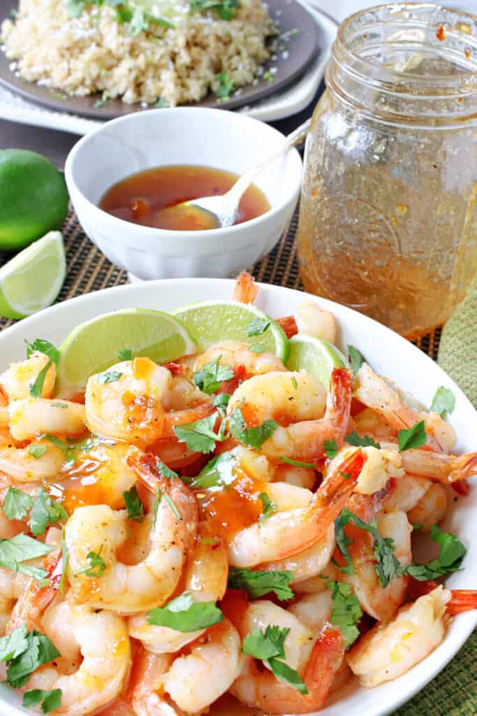 A vertical closeup of Whiskey Glazed Roasted Shrimp in the foreground along with a bowl of rice and a a jar of glaze in the background.