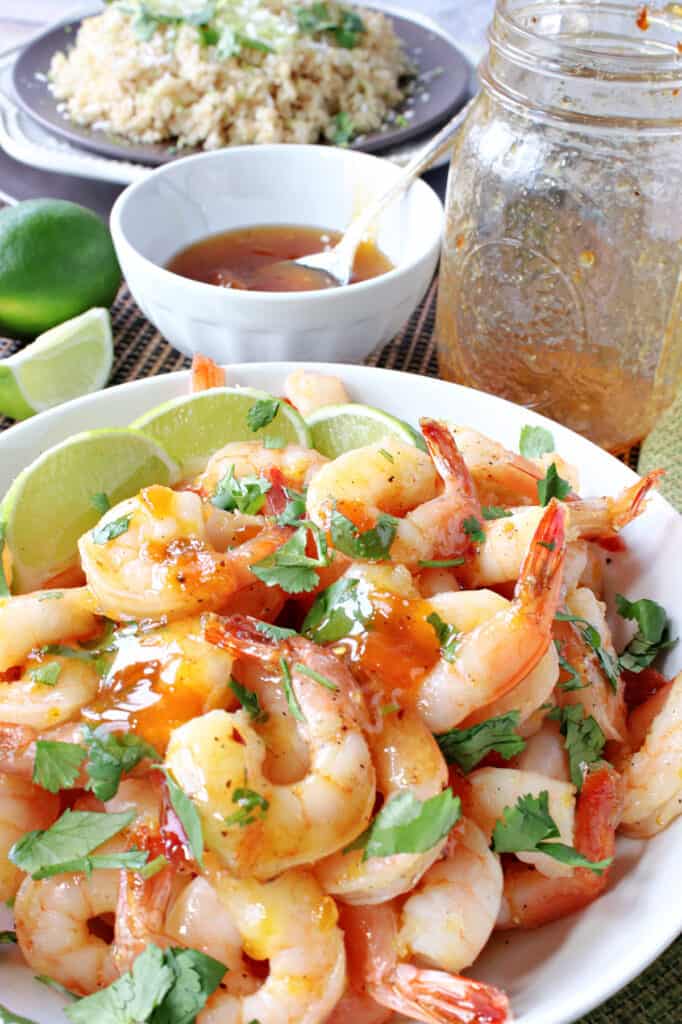 A vertical closeup photo of Whiskey Glazed Roasted Shrimp in a white bowl along with lime slices and cilantro.