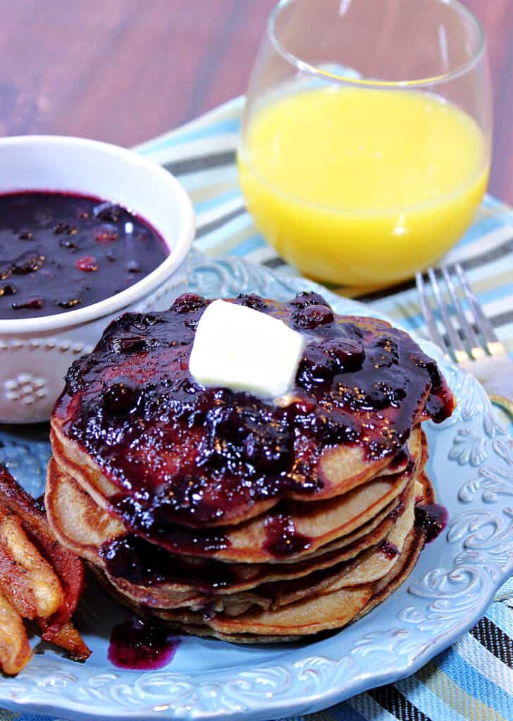 A stack of Whole Wheat Peanut Butter Pancakes topped with Blueberry Maple Syrup and a glass of orange juice in the background.