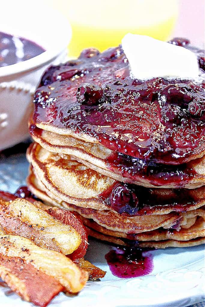 A closeup vertical image of Whole Wheat Peanut Butter Pancakes along with Blueberry Maple Syrup and bacon on a plate.