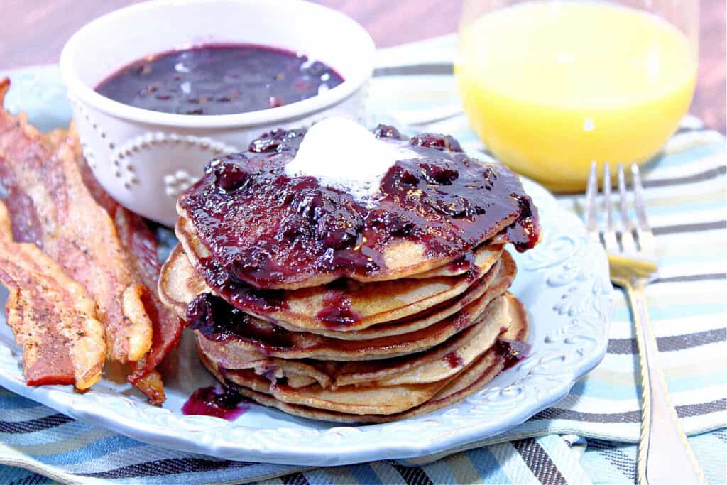 A stack of Whole Wheat Peanut Butter Pancakes on a blue plate with bacon and a topping of Blueberry Maple Syrup.