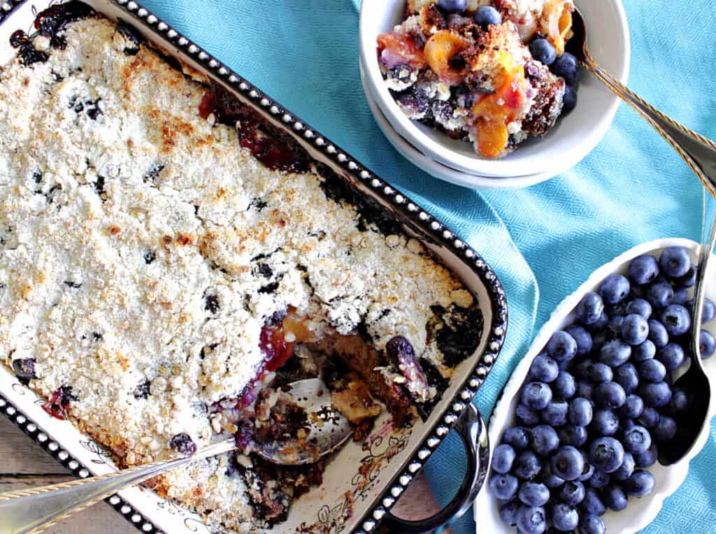 A direct overhead photo of a baking dish filled with Blueberry Peach Crisp along with a bowl of fresh blueberries and a few spoons.