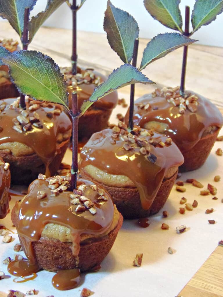 A vertical closeup of caramel topped Caramel Apple Cupcakes with stems, leaves, and chopped pecans.
