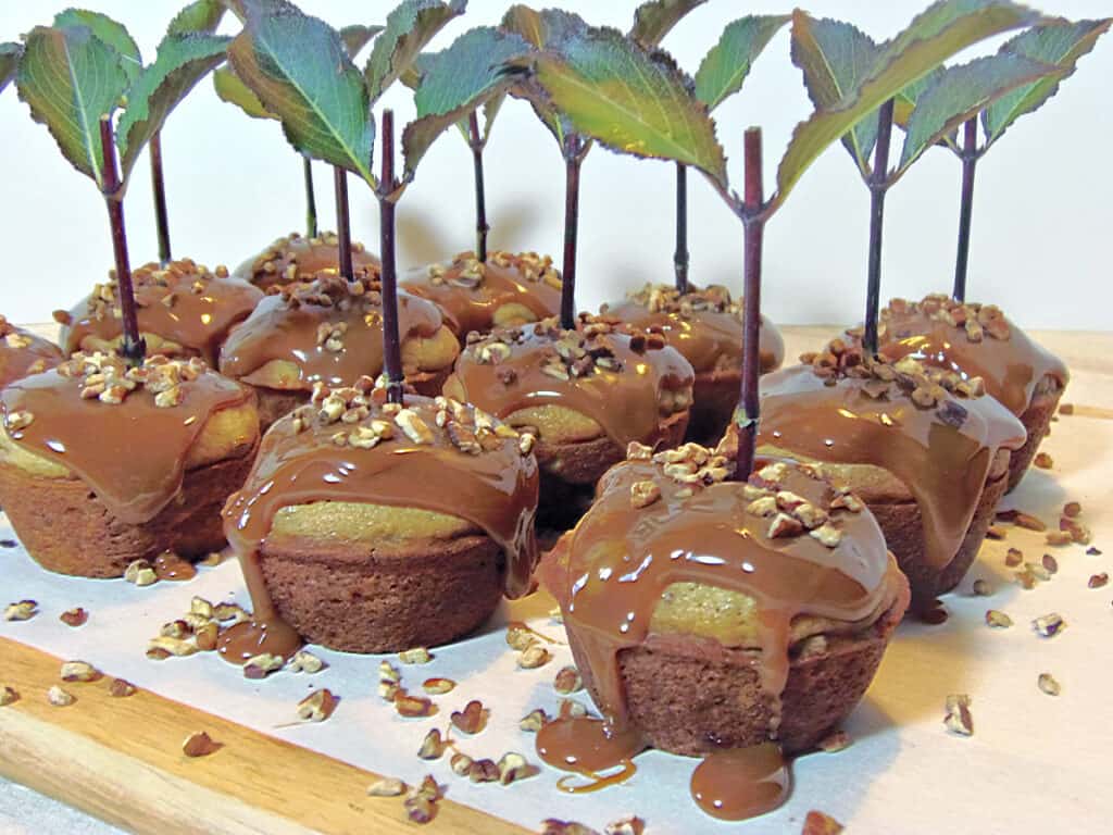 A lineup of Caramel Apple Cupcakes on a wooden board with dripping caramel, and chopped pecans.