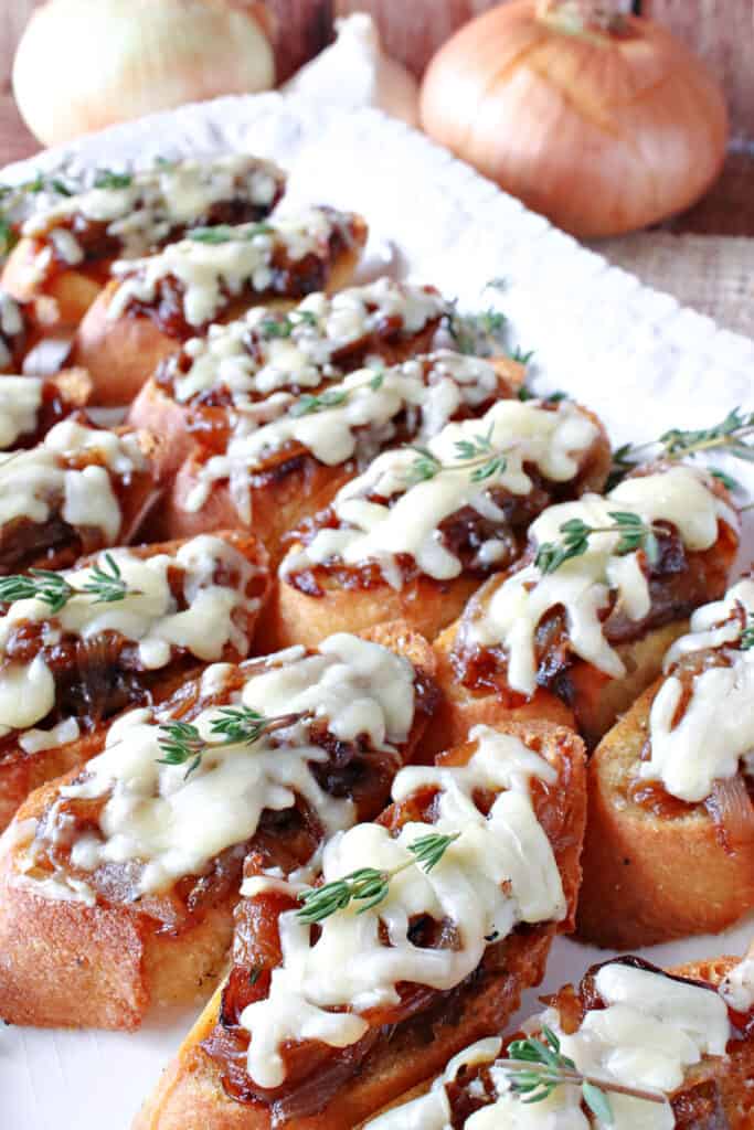 A vertical closeup of a platter of French Onion Crostini along with melted cheese, fresh thyme, with  onions and garlic in the background.